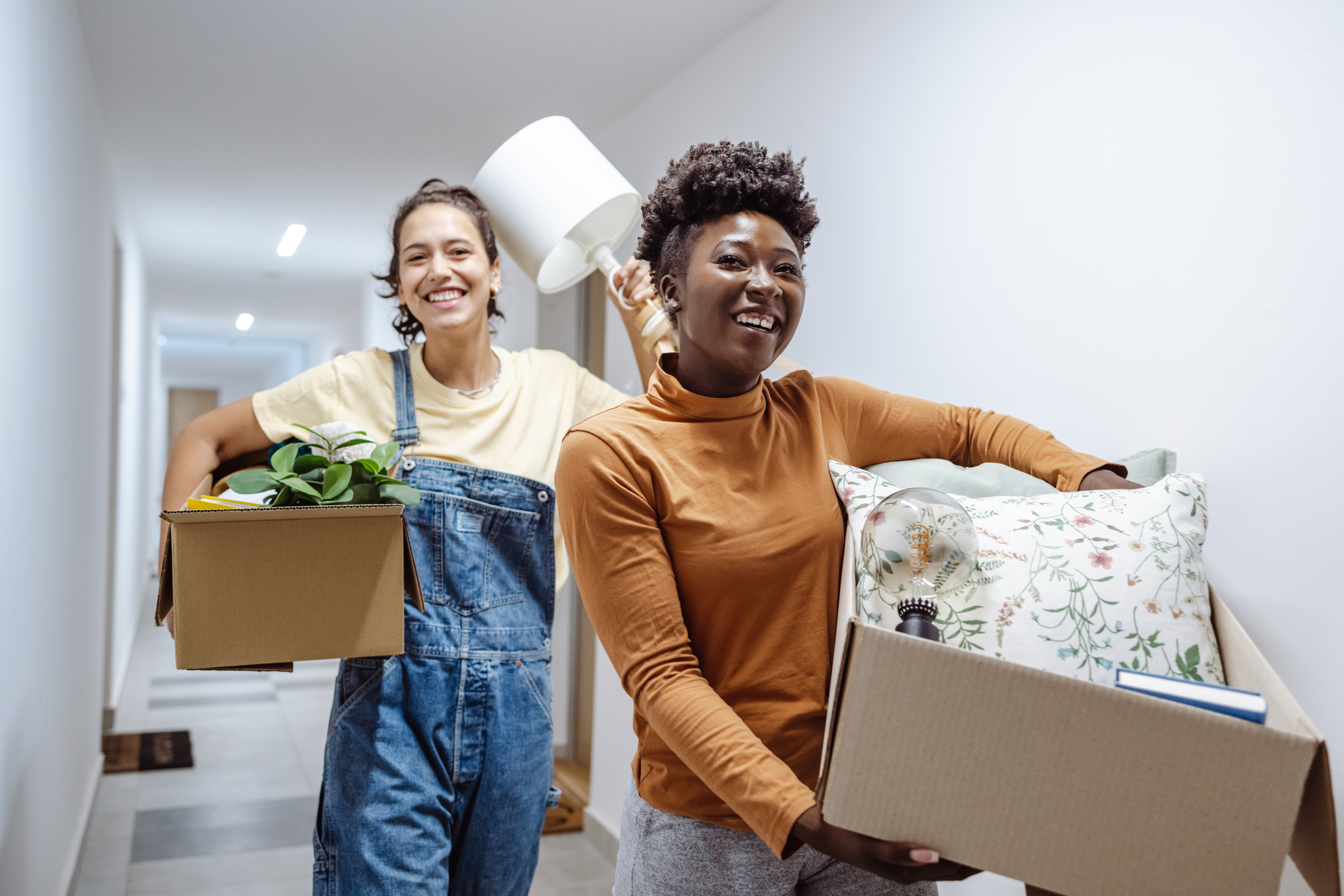 Two college aged women walking down a dorm hallway with full boxes during move out